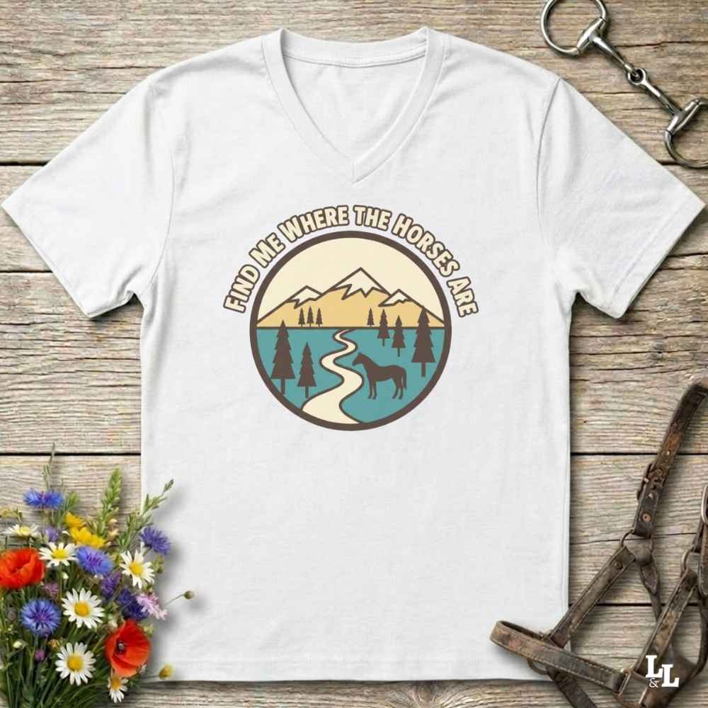 A white V-neck t-shirt with a retro mountain landscape graphic and the text 'FIND ME WHERE THE HORSES ARE' laid flat on a rustic wooden background with equestrian and floral props.