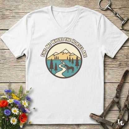 A white V-neck t-shirt with a retro mountain landscape graphic and the text 'FIND ME WHERE THE HORSES ARE' laid flat on a rustic wooden background with equestrian and floral props.