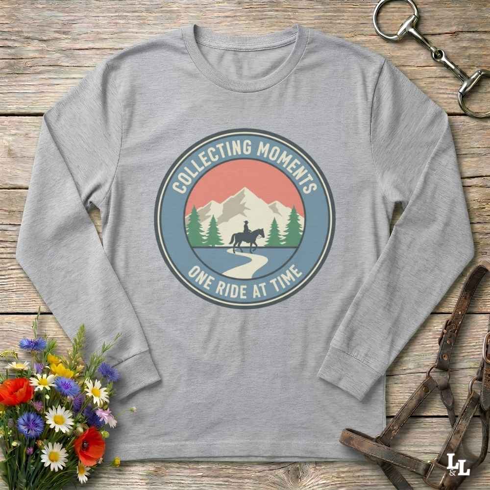 A heather gray long-sleeve t-shirt with a circular retro graphic on the chest that reads 'COLLECTING MOMENTS' and 'ONE RIDE AT TIME' around an illustration of mountains, pine trees, and a horse rider. The shirt is displayed flat on a rustic wooden background with equestrian-themed props like a leather bridle and a bouquet of wildflowers.