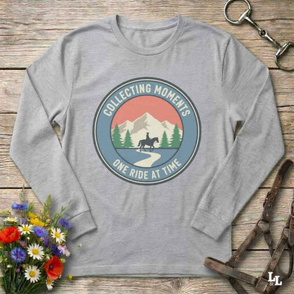 A heather gray long-sleeve t-shirt with a circular retro graphic on the chest that reads 'COLLECTING MOMENTS' and 'ONE RIDE AT TIME' around an illustration of mountains, pine trees, and a horse rider. The shirt is displayed flat on a rustic wooden background with equestrian-themed props like a leather bridle and a bouquet of wildflowers.