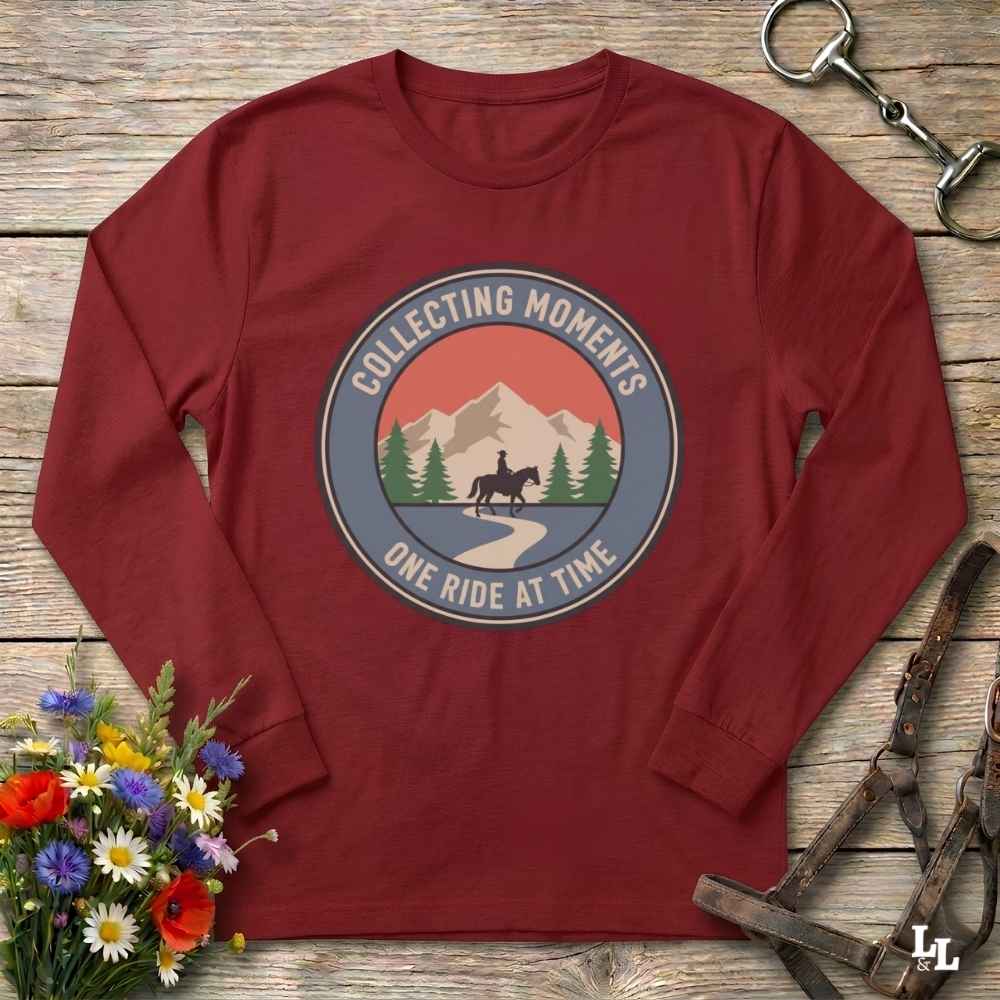 A long-sleeve crewneck t-shirt in maroon red, laid flat on a rustic wooden background. The shirt features a circular graphic print in the center with a horse rider on a path between mountains and pine trees, captioned 'COLLECTING MOMENTS' and 'ONE RIDE AT TIME'.