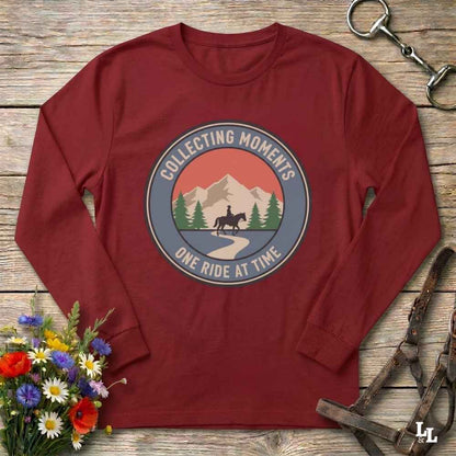 A long-sleeve crewneck t-shirt in maroon red, laid flat on a rustic wooden background. The shirt features a circular graphic print in the center with a horse rider on a path between mountains and pine trees, captioned 'COLLECTING MOMENTS' and 'ONE RIDE AT TIME'.