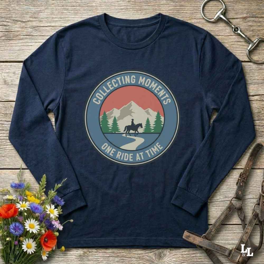 A navy blue long-sleeve t-shirt with a circular graphic on the chest that reads 'COLLECTING MOMENTS' and 'ONE RIDE AT TIME' around an illustration of mountains, trees, and a horse rider. The shirt is displayed in a flat-lay arrangement on a rustic wooden background with equestrian accessories and flowers.