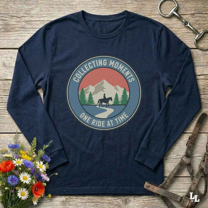 A navy blue long-sleeve t-shirt with a circular graphic on the chest that reads 'COLLECTING MOMENTS' and 'ONE RIDE AT TIME' around an illustration of mountains, trees, and a horse rider. The shirt is displayed in a flat-lay arrangement on a rustic wooden background with equestrian accessories and flowers.
