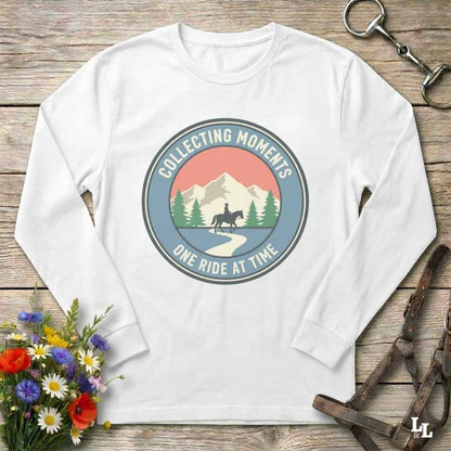 A white long-sleeve crewneck t-shirt with a circular retro graphic on the chest that reads 'COLLECTING MOMENTS' and 'ONE RIDE AT TIME' around an illustration of a horse rider, mountains, and pine trees. The shirt is displayed in a flat-lay arrangement on a rustic wooden background, styled with a small bouquet of wildflowers, a leather bridle, and a metal bit.