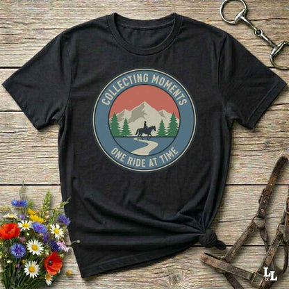 A black short-sleeve graphic T-shirt laid flat on a rustic wooden surface, styled with flowers, a horse bit, and leather tack. The shirt has a circular retro-style mountain landscape print with a horse rider silhouette and the text 'COLLECTING MOMENTS' and 'ONE RIDE AT TIME'.