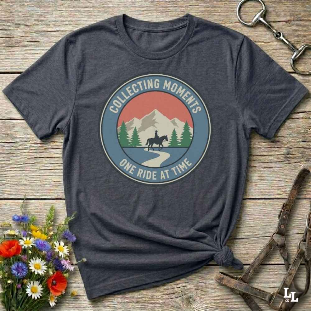 A dark heather gray graphic t-shirt laid flat on a rustic wooden surface, styled with a bouquet of wildflowers, a horse bit, and leather tack. The t-shirt features a circular retro graphic with a horse rider, mountains, and the text 'COLLECTING MOMENTS' and 'ONE RIDE AT TIME'.