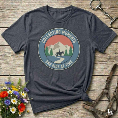 A dark heather gray graphic t-shirt laid flat on a rustic wooden surface, styled with a bouquet of wildflowers, a horse bit, and leather tack. The t-shirt features a circular retro graphic with a horse rider, mountains, and the text 'COLLECTING MOMENTS' and 'ONE RIDE AT TIME'.
