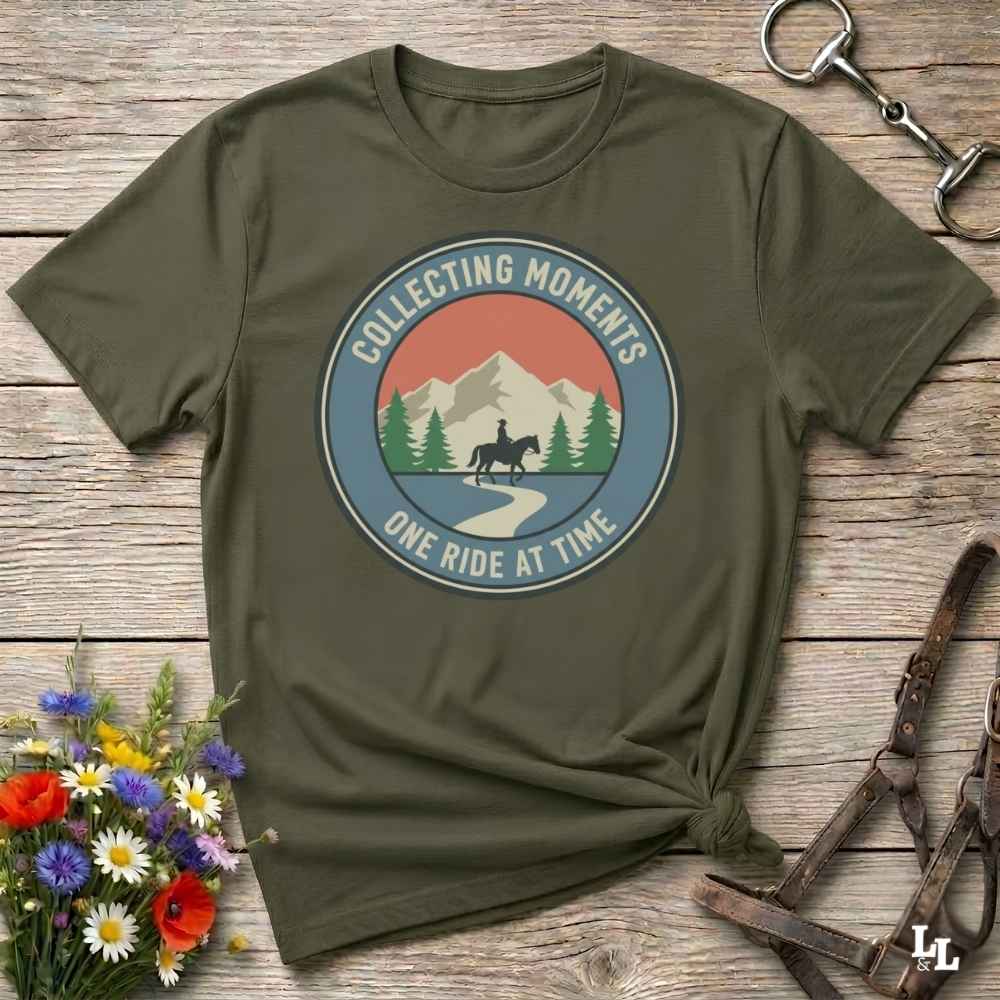 An olive green short-sleeve t-shirt with a circular graphic on the chest that reads 'COLLECTING MOMENTS' and 'ONE RIDE AT TIME' around an illustration of mountains, pine trees, and a horse rider. The shirt is styled in a flat-lay on a rustic wooden surface with a horse bit, leather bridle, and a small bouquet of wildflowers.