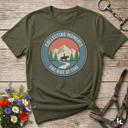 An olive green short-sleeve t-shirt with a circular graphic on the chest that reads 'COLLECTING MOMENTS' and 'ONE RIDE AT TIME' around an illustration of mountains, pine trees, and a horse rider. The shirt is styled in a flat-lay on a rustic wooden surface with a horse bit, leather bridle, and a small bouquet of wildflowers.