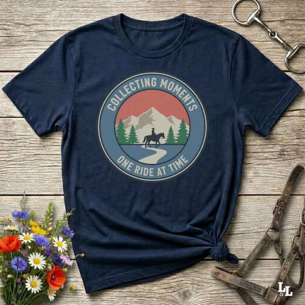 A navy blue short-sleeve t-shirt with a circular graphic on the chest that reads 'Collecting Moments One Ride at Time' around an illustration of mountains, trees, and a horse rider. The shirt is styled in a flat-lay on a rustic wooden background with equestrian accessories and flowers.