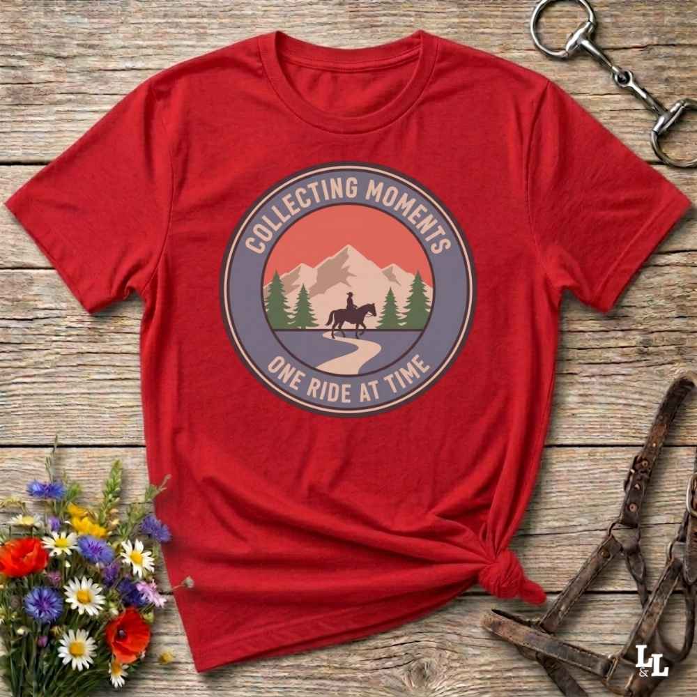 A red short-sleeve graphic T-shirt laid flat on a rustic wooden surface, with a circular mountain-and-horse print reading 'Collecting Moments' and 'One Ride At Time.' Decorative flowers and horse tack are placed around it as styling props.