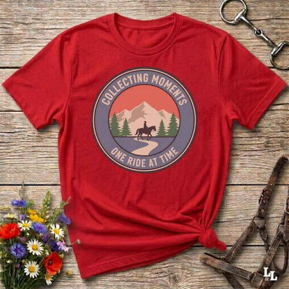 A red short-sleeve graphic T-shirt laid flat on a rustic wooden surface, with a circular mountain-and-horse print reading 'Collecting Moments' and 'One Ride At Time.' Decorative flowers and horse tack are placed around it as styling props.