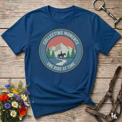 A navy blue short-sleeve t-shirt with a circular graphic on the chest reading 'COLLECTING MOMENTS' and 'ONE RIDE AT TIME' around an illustration of mountains, trees, a horse, and a rider. The shirt is styled in a flat lay on a rustic wooden surface with flowers and leather tack nearby.