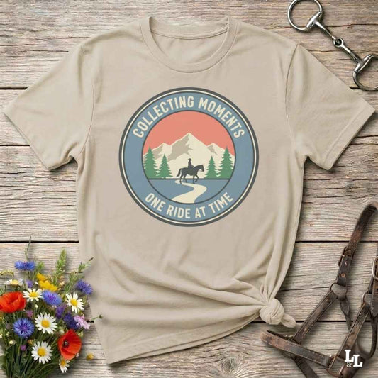 A beige graphic t-shirt with a circular mountain and horse design, styled on a rustic wooden background with flowers and leather straps.