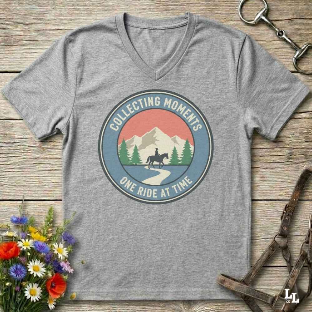 A heather gray v-neck t-shirt with a circular retro graphic on the chest reading 'Collecting Moments One Ride at Time' around a mountain and rider illustration. The shirt is displayed flat on a rustic wooden background with equestrian and floral props.