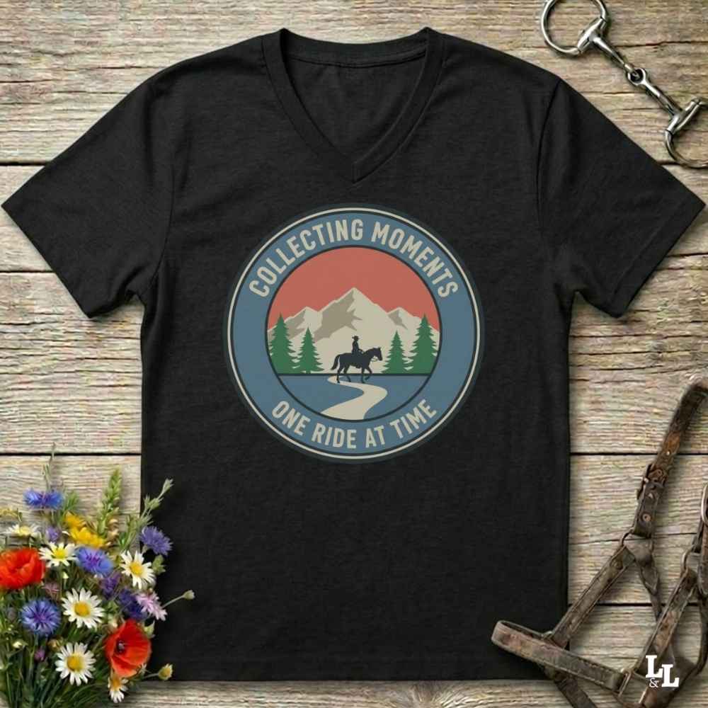 A black V-neck t-shirt with a retro circular graphic that reads 'COLLECTING MOMENTS ONE RIDE AT TIME' around an illustration of mountains, pine trees, and a horse rider. The shirt is laid flat on a rustic wooden background with equestrian accessories and flowers as props.