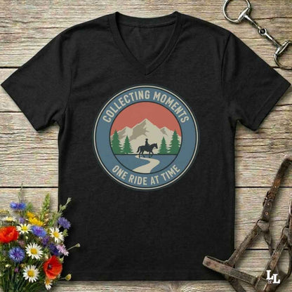 A black V-neck t-shirt with a retro circular graphic that reads 'COLLECTING MOMENTS ONE RIDE AT TIME' around an illustration of mountains, pine trees, and a horse rider. The shirt is laid flat on a rustic wooden background with equestrian accessories and flowers as props.