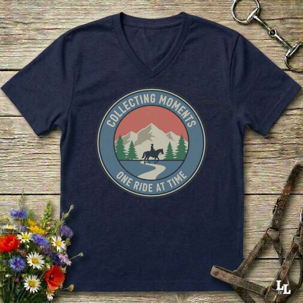 A navy blue V-neck t-shirt with a circular graphic print featuring a mountain, trees, and a horse rider, with the text 'COLLECTING MOMENTS' and 'ONE RIDE AT TIME'. The shirt is displayed on a rustic wooden background with equestrian accessories and flowers.