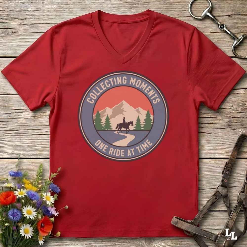 A red V-neck t-shirt laid flat on a rustic wooden surface, featuring a circular graphic with a mountain, trees, and a horse rider, with the text 'COLLECTING MOMENTS' and 'ONE RIDE AT TIME'. The shirt is styled with equestrian tack and a small bouquet of wildflowers.