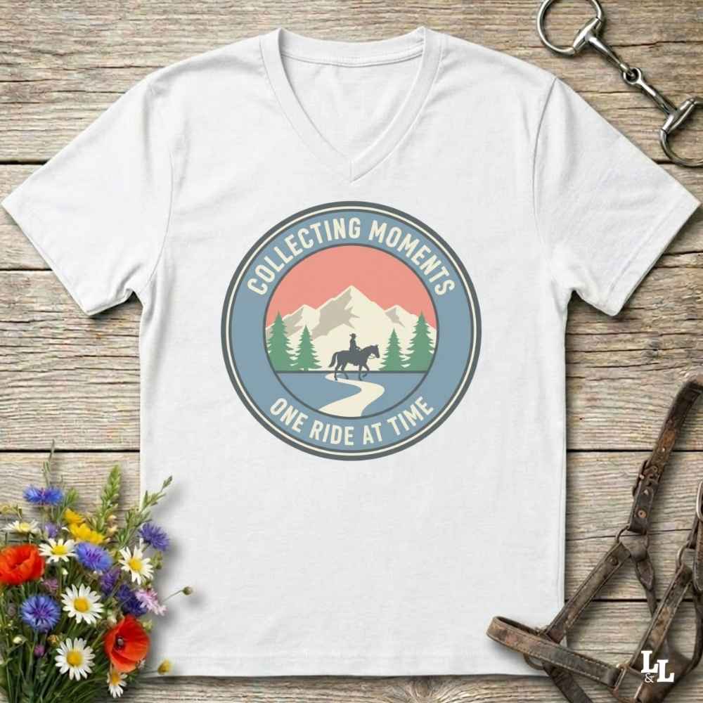 A white V-neck t-shirt laid flat on a rustic wooden surface, featuring a circular retro graphic with mountains, trees, a horse rider, and the text 'COLLECTING MOMENTS ONE RIDE AT TIME.' Decorative flowers and horse tack are placed around the shirt.