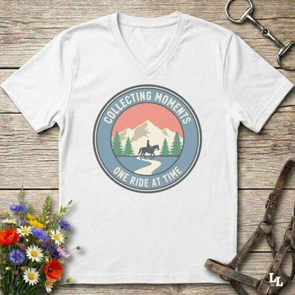 A white V-neck t-shirt laid flat on a rustic wooden surface, featuring a circular retro graphic with mountains, trees, a horse rider, and the text 'COLLECTING MOMENTS ONE RIDE AT TIME.' Decorative flowers and horse tack are placed around the shirt.