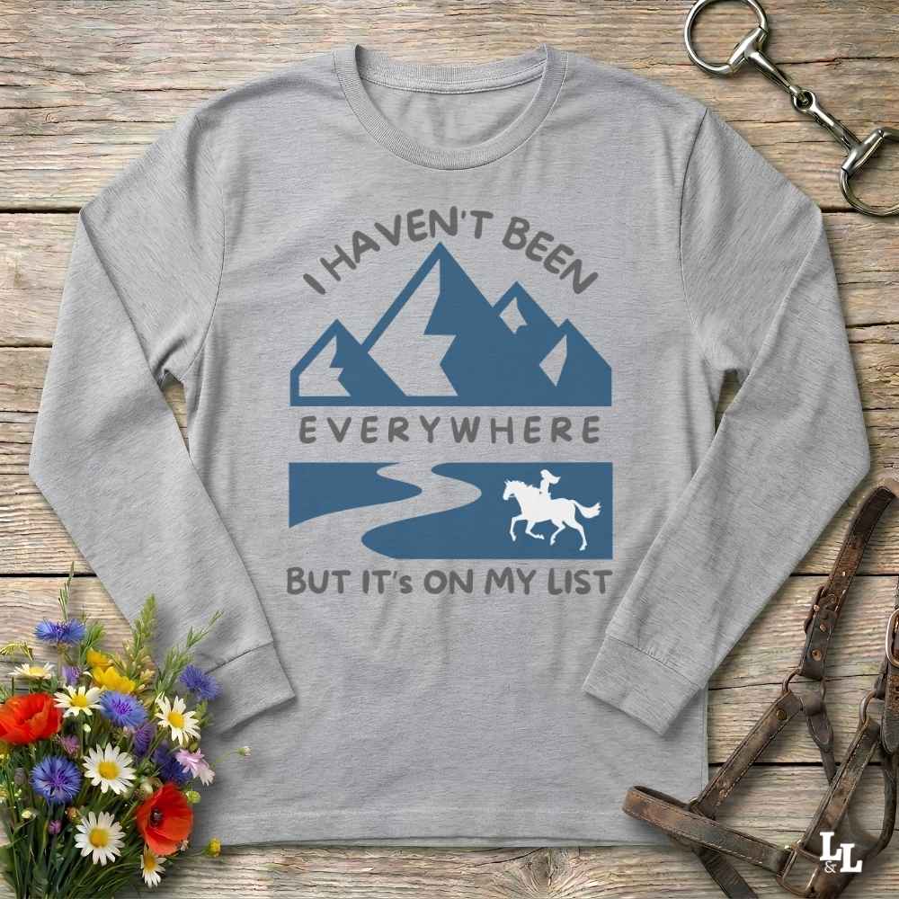 A light heather gray long-sleeve t-shirt with a blue and gray graphic print of mountains, a river, and a horse rider, laid flat on a rustic wooden surface with decorative flowers and leather gear.