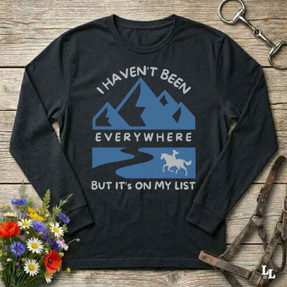 A black long-sleeve t-shirt with a blue and white graphic print featuring mountains, a river, a horse, and the text 'I HAVEN'T BEEN EVERYWHERE BUT IT'S ON MY LIST'. The shirt is displayed flat on a rustic wooden background with equestrian accessories and flowers.