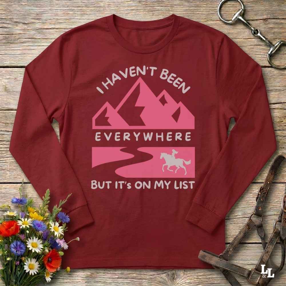 A long-sleeve crewneck t-shirt in maroon red, laid flat on a rustic wooden background. The shirt features a pink and white graphic with mountains, a winding road, and a horse silhouette, accompanied by the text 'I HAVEN'T BEEN EVERYWHERE BUT IT'S ON MY LIST'.