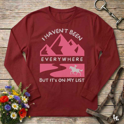 A long-sleeve crewneck t-shirt in maroon red, laid flat on a rustic wooden background. The shirt features a pink and white graphic with mountains, a winding road, and a horse silhouette, accompanied by the text 'I HAVEN'T BEEN EVERYWHERE BUT IT'S ON MY LIST'.