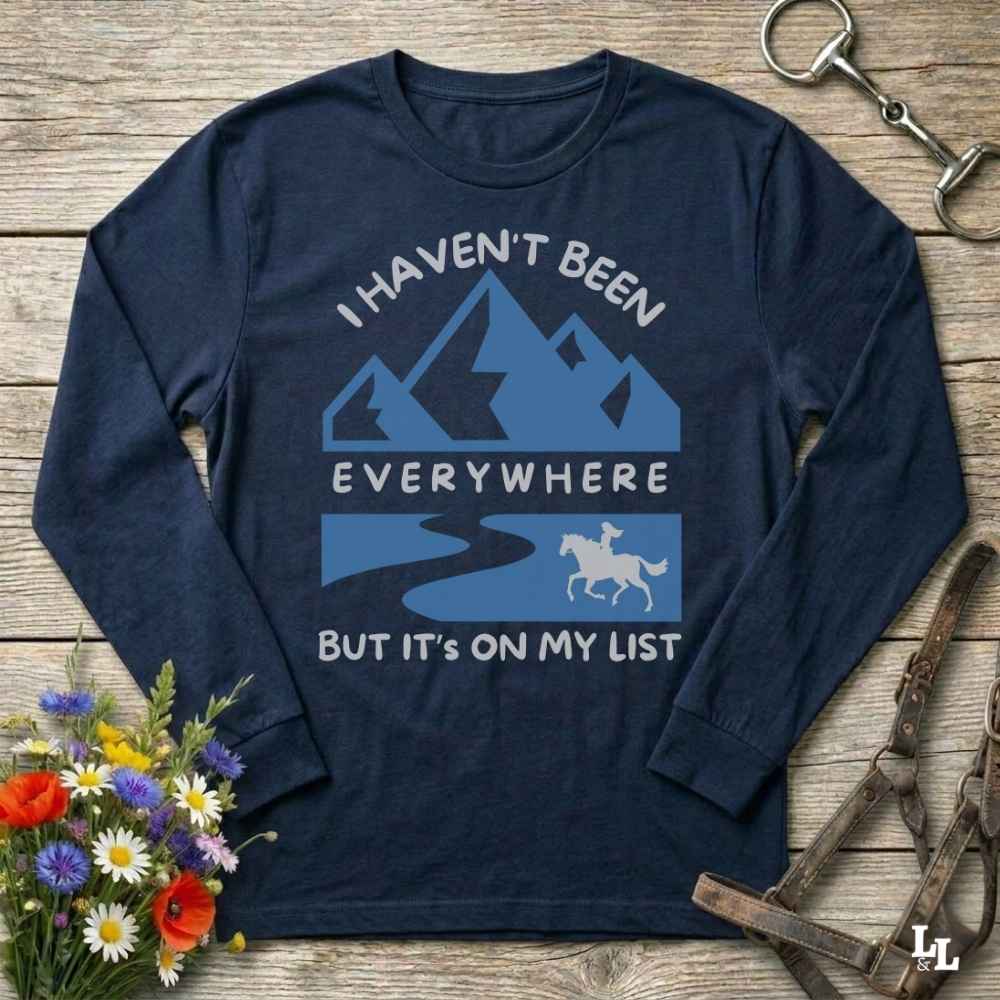 A navy blue long-sleeve t-shirt with a graphic print of mountains, a river, and a horse rider, accompanied by the text 'I HAVEN'T BEEN EVERYWHERE BUT IT'S ON MY LIST'. The shirt is displayed flat on a rustic wooden background with decorative flowers and horse tack.