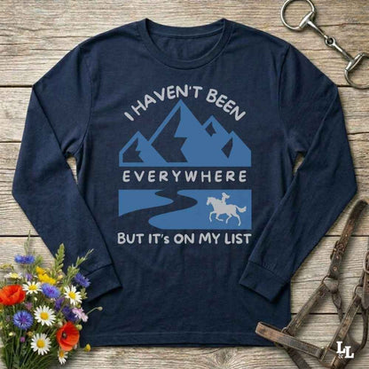 A navy blue long-sleeve t-shirt with a graphic print of mountains, a river, and a horse rider, accompanied by the text 'I HAVEN'T BEEN EVERYWHERE BUT IT'S ON MY LIST'. The shirt is displayed flat on a rustic wooden background with decorative flowers and horse tack.
