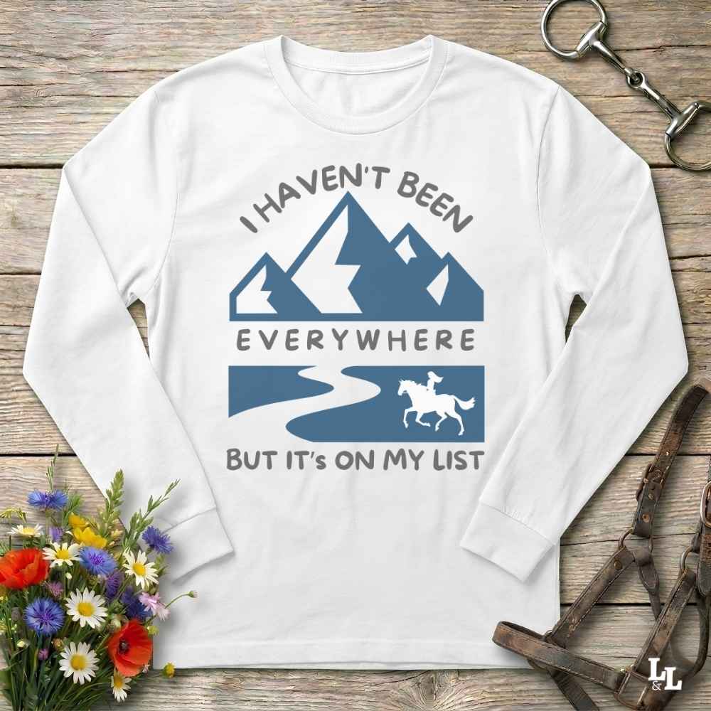 A white long-sleeve t-shirt with a blue and gray graphic print featuring mountains, a river, and a horse rider, laid flat on a rustic wooden background with decorative flowers and leather gear.