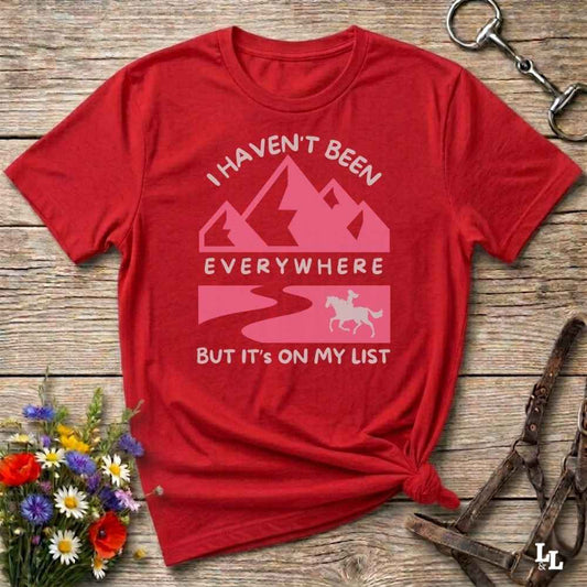 A red short-sleeve graphic T-shirt laid flat on a rustic wooden surface, with a pink mountain and winding road design and the text 'I HAVEN'T BEEN EVERYWHERE BUT IT'S ON MY LIST.' Decorative flowers and horse tack are placed around it as styling props.