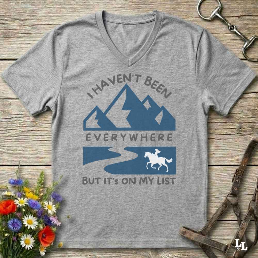 A heather gray V-neck t-shirt with a blue and white graphic print that reads 'I HAVEN'T BEEN EVERYWHERE BUT IT'S ON MY LIST' with mountain, river, and horse illustrations. The shirt is displayed flat on a rustic wooden background with decorative flowers and horse tack.