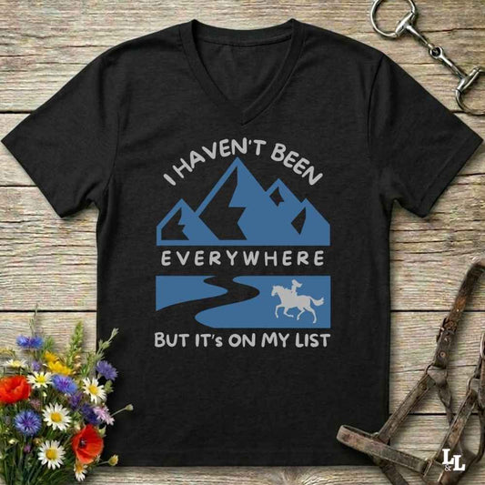 A black V-neck t-shirt with a blue and white graphic print reading 'I HAVEN'T BEEN EVERYWHERE BUT IT'S ON MY LIST' with mountain, river, and horse illustrations, displayed on a rustic wooden background with flowers and horse tack props.