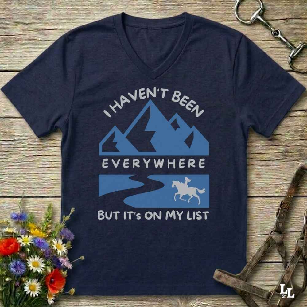 A navy blue V-neck t-shirt with a light blue and white graphic print that reads 'I HAVEN'T BEEN EVERYWHERE BUT IT'S ON MY LIST' with mountain, river, and horse illustrations. The shirt is displayed in a flat-lay arrangement on a rustic wooden background with decorative flowers and horse tack.