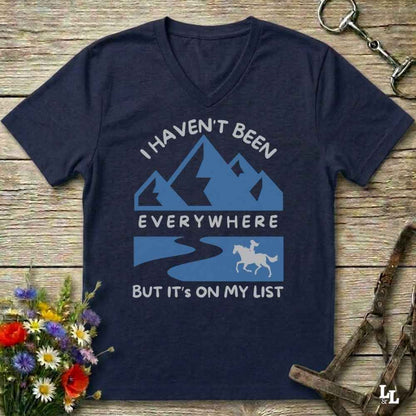 A navy blue V-neck t-shirt with a light blue and white graphic print that reads 'I HAVEN'T BEEN EVERYWHERE BUT IT'S ON MY LIST' with mountain, river, and horse illustrations. The shirt is displayed in a flat-lay arrangement on a rustic wooden background with decorative flowers and horse tack.