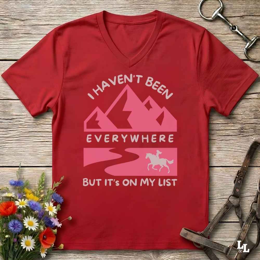 A red V-neck t-shirt with a pink and white graphic print that reads 'I HAVEN'T BEEN EVERYWHERE BUT IT'S ON MY LIST' with mountain, road, and horse illustrations. The shirt is displayed flat on a rustic wooden background with equestrian tack and flowers as props.