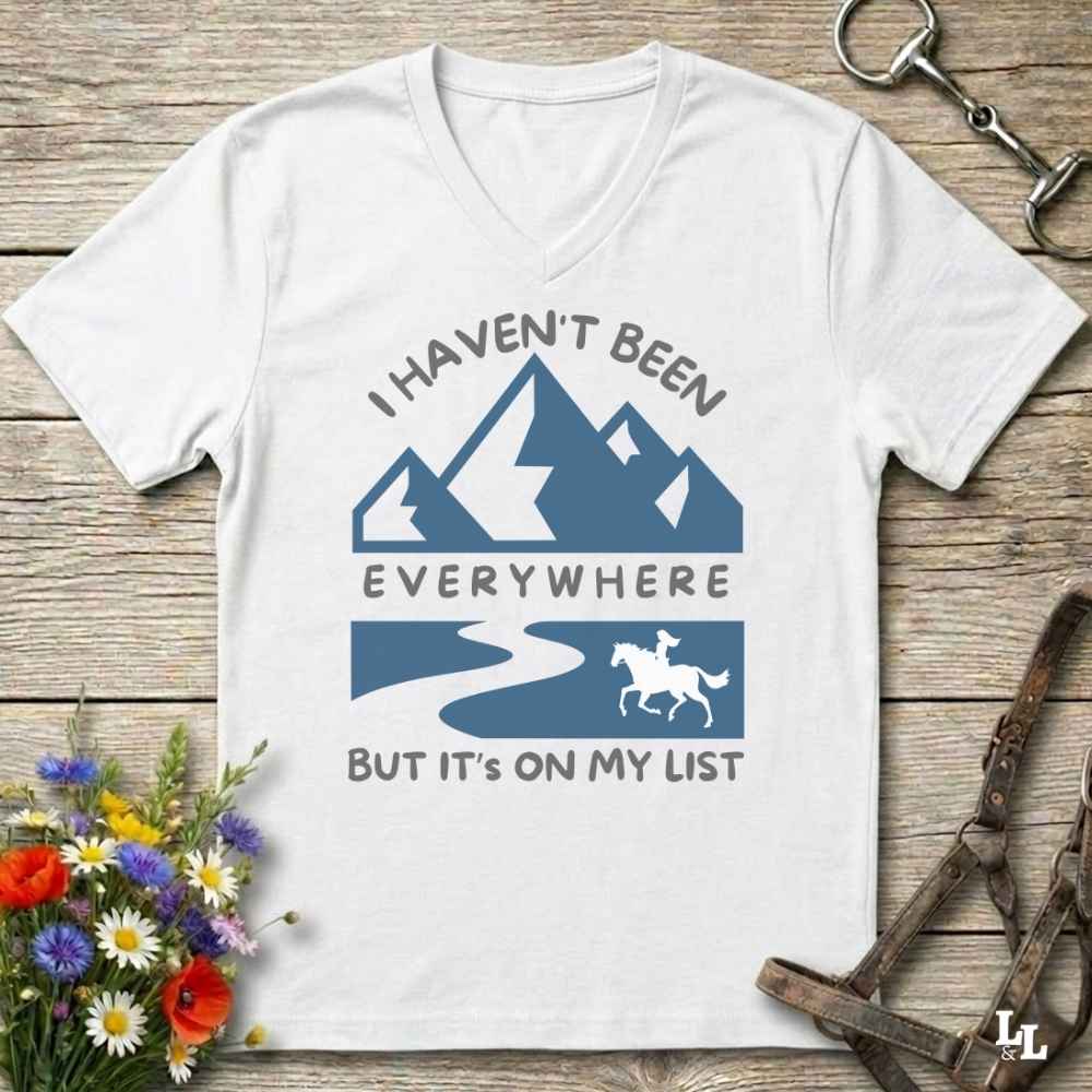 A white V-neck t-shirt laid flat on a rustic wooden surface, featuring a blue graphic of mountains, a winding road, a horse, and the text 'I HAVEN'T BEEN EVERYWHERE BUT IT'S ON MY LIST'. The shirt is styled with equestrian and floral props.