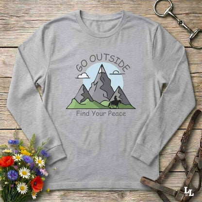 A heather gray long-sleeve t-shirt with a graphic of mountains, a horse, and the text 'GO OUTSIDE Find Your Peace', laid flat on a rustic wooden surface with decorative flowers and leather gear around it.