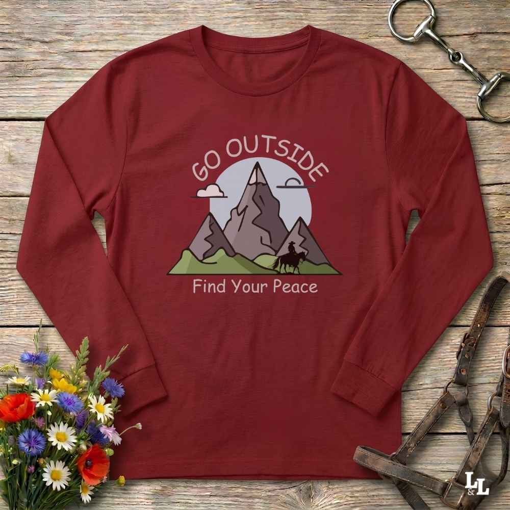 A long-sleeve crewneck t-shirt in maroon red, laid flat on a rustic wooden surface. The shirt features a graphic of mountains, a horse, and a sun with the text 'GO OUTSIDE Find Your Peace'.