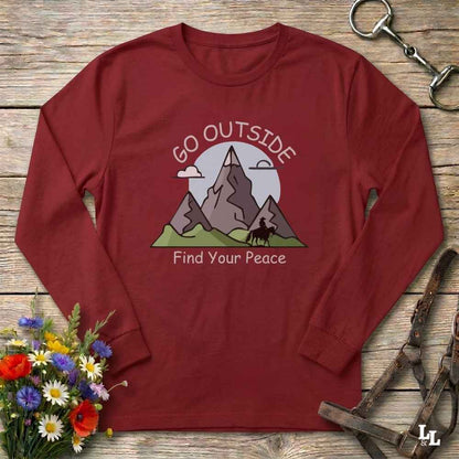 A long-sleeve crewneck t-shirt in maroon red, laid flat on a rustic wooden surface. The shirt features a graphic of mountains, a horse, and a sun with the text 'GO OUTSIDE Find Your Peace'.