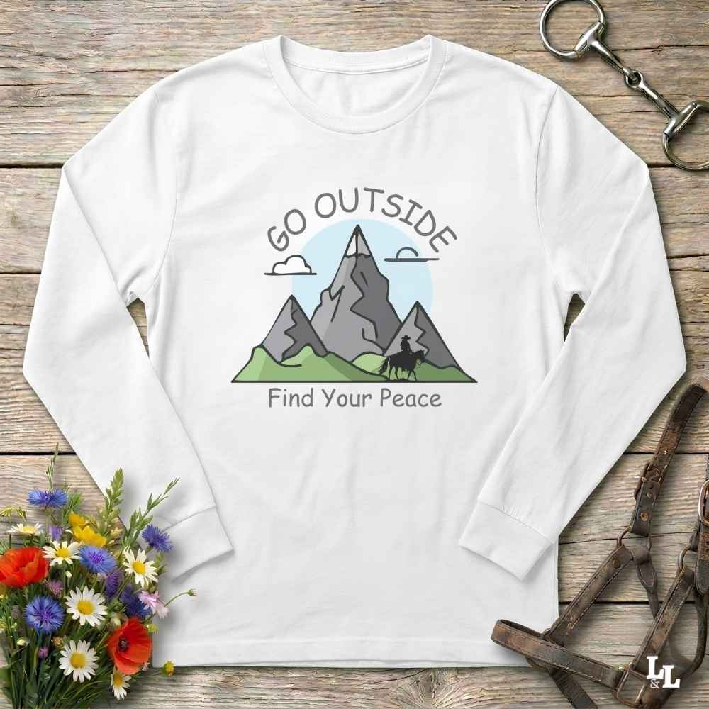A white long-sleeve t-shirt with a graphic of mountains, a horse, and the text 'GO OUTSIDE Find Your Peace', laid flat on a rustic wooden background with equestrian accessories and flowers.