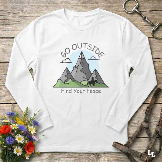 A white long-sleeve t-shirt with a graphic of mountains, a horse, and the text 'GO OUTSIDE Find Your Peace', laid flat on a rustic wooden background with equestrian accessories and flowers.