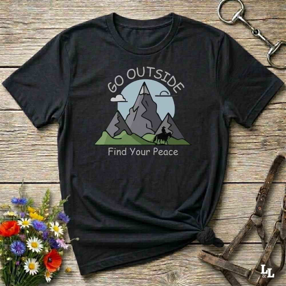 A black short-sleeve graphic T-shirt laid flat on a rustic wooden surface, styled with flowers, a horse bit, and leather straps. The shirt has a mountain landscape print with the words 'GO OUTSIDE Find Your Peace' in white.