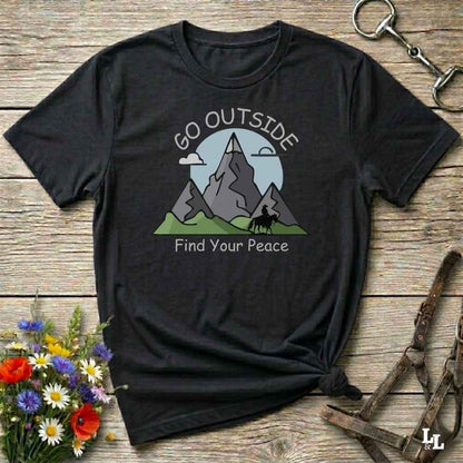 A black short-sleeve graphic T-shirt laid flat on a rustic wooden surface, styled with flowers, a horse bit, and leather straps. The shirt has a mountain landscape print with the words 'GO OUTSIDE Find Your Peace' in white.