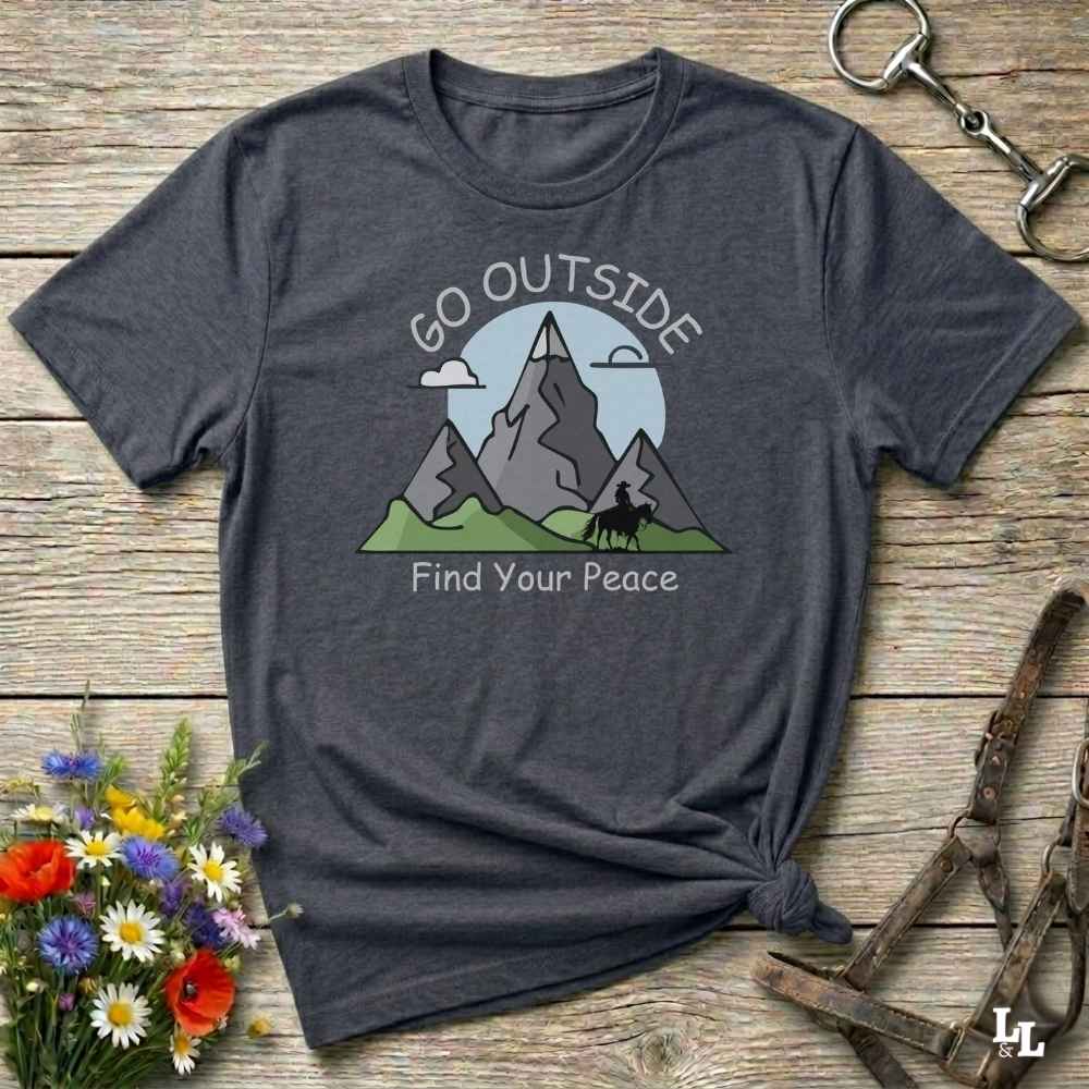 A dark heather gray t-shirt with a graphic of mountains, a tree, and the text 'GO OUTSIDE Find Your Peace', styled on a rustic wooden background with a horse bit and flowers.