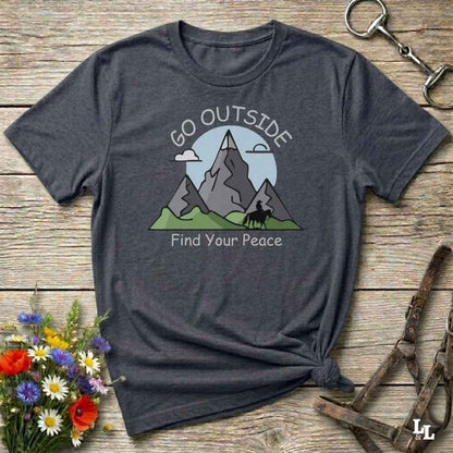 A dark heather gray t-shirt with a graphic of mountains, a tree, and the text 'GO OUTSIDE Find Your Peace', styled on a rustic wooden background with a horse bit and flowers.
