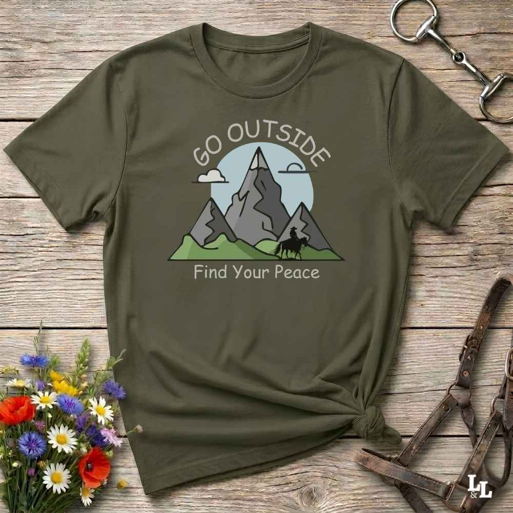 An olive green graphic t-shirt laid flat on a rustic wooden surface, styled with a small bouquet of flowers, a metal horse bit, and leather straps. The t-shirt features a graphic of gray mountains, green hills, and a light blue sun with the text 'GO OUTSIDE Find Your Peace'.